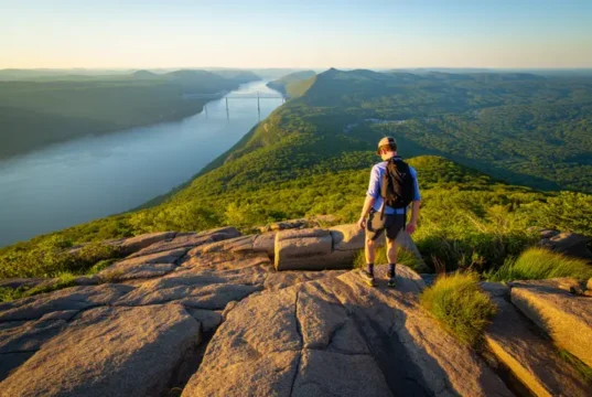 Major Welch Trail Bear Mountain: An Expert Field Guide A hiker on a rock scramble on the Major Welch Trail, looking at the panoramic view of the Hudson River from Bear Mountain at sunset.