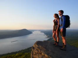 Two hikers mastering the tough Breakneck Ridge trail in New York, looking at the panoramic Hudson River view at sunset.