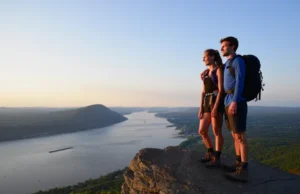 Mastering Breakneck Ridge: NY’s Toughest Day Hike Two hikers mastering the tough Breakneck Ridge trail in New York, looking at the panoramic Hudson River view at sunset.
