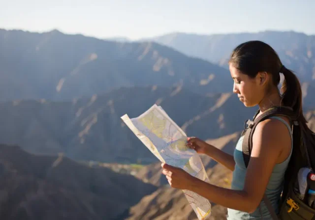 A hiker stands at a Southern California mountain overlook at sunrise, thoughtfully studying a map to plan her route for a skill-building hike.