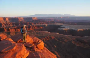 A Hiker’s Guide to St. George: 12 Trails Analyzed A hiker gazes out at a sweeping sunrise vista of canyons and mountains from a red rock overlook near St. George, Utah.