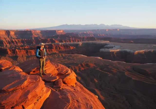 A hiker gazes out at a sweeping sunrise vista of canyons and mountains from a red rock overlook near St. George, Utah.