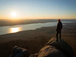 A hiker stands on the rocky summit of Turkey Mountain Nature Preserve, looking out at a panoramic sunset view of the Croton Reservoir and the distant NYC skyline.