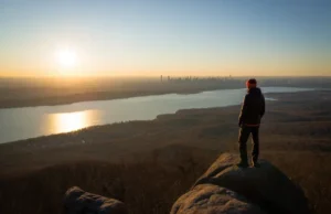 A Hiker’s Guide to Turkey Mountain Nature Preserve A hiker stands on the rocky summit of Turkey Mountain Nature Preserve, looking out at a panoramic sunset view of the Croton Reservoir and the distant NYC skyline.