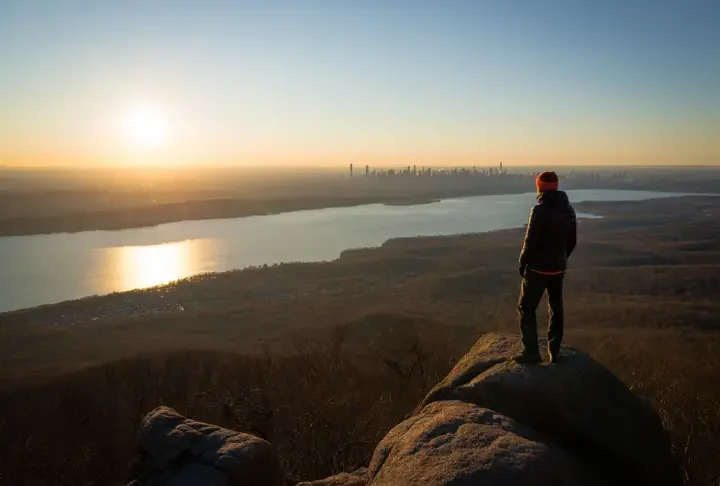 A hiker stands on the rocky summit of Turkey Mountain Nature Preserve, looking out at a panoramic sunset view of the Croton Reservoir and the distant NYC skyline.
