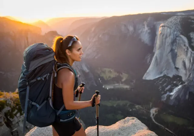 A skilled female hiker with a backpack looks out over a spectacular mountain valley at sunrise, representing the rewarding challenge of USA bucket list hikes.