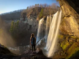 A wide-angle view of Verkeerder Kill Falls cascading down a dramatic cliff in the Shawangunk Mountains, with a hiker looking on from a sunlit, rocky overlook during golden hour.