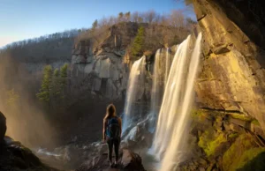 Field-Tested Guide: Verkeerder Kill Falls Hike (NY) A wide-angle view of Verkeerder Kill Falls cascading down a dramatic cliff in the Shawangunk Mountains, with a hiker looking on from a sunlit, rocky overlook during golden hour.