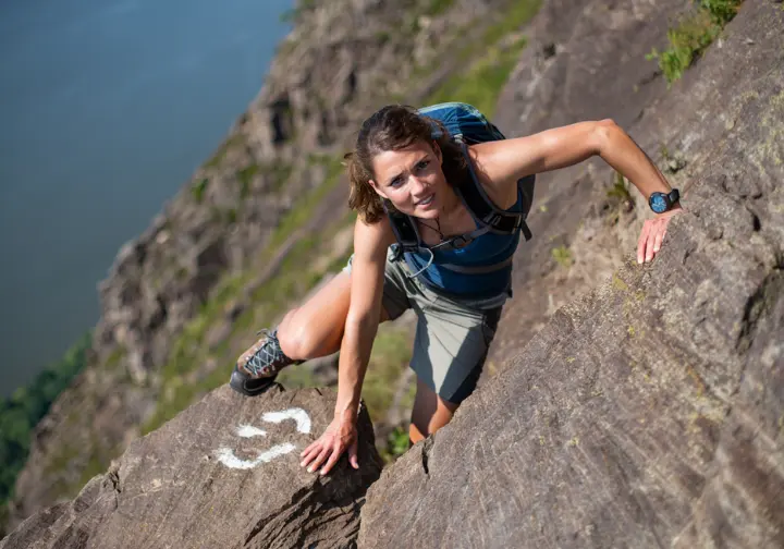 A hiker carefully scrambles up the steep, white-blazed rock face of the Breakneck Ridge trail, demonstrating the challenging climb.