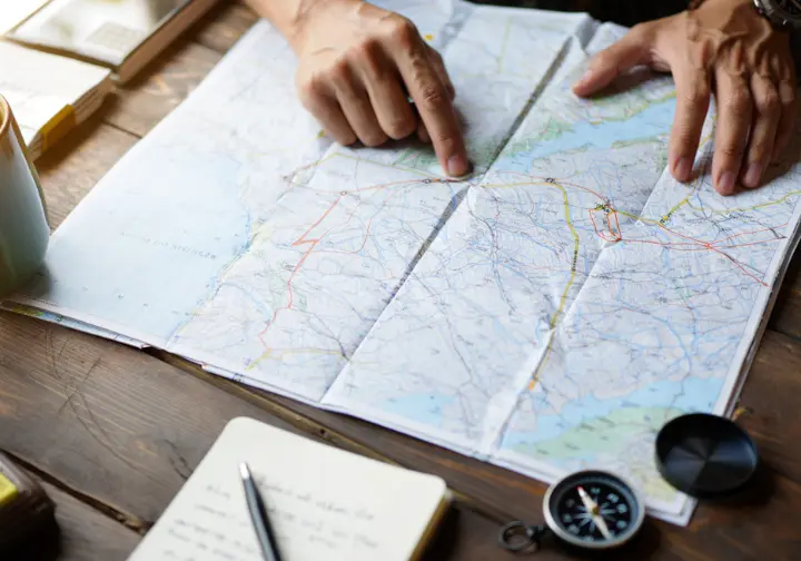 A hiker's hands carefully trace a route on a topographical map, surrounded by a compass and notebook, illustrating the planning that goes beyond a simple trail list.