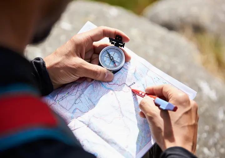 A close-up of a hiker's hands using a compass and a topographic map, illustrating the philosophy of approaching hiking as a skill-based practice.