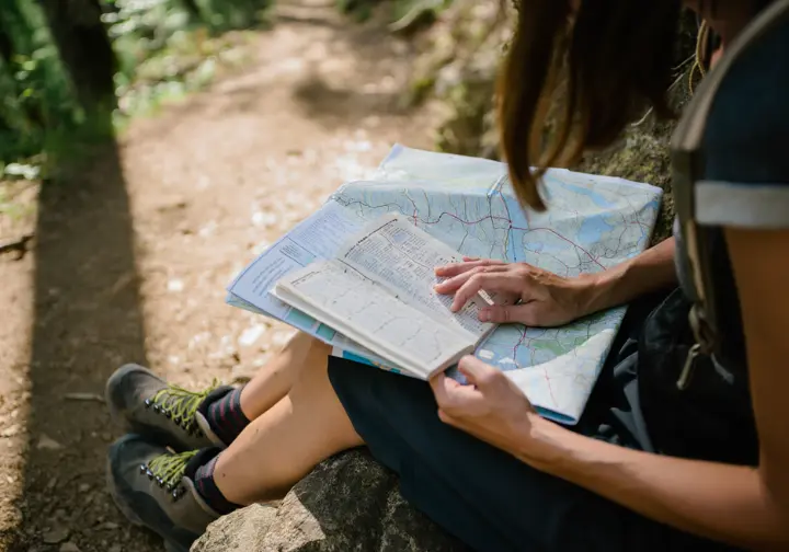 A hiker sits on a rock carefully studying a topographic map and a guidebook to understand how bucket list hikes are ranked by skill level.