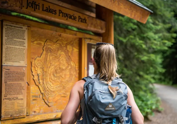 A hiker studies a wooden map and information sign at the Joffre Lakes trailhead before starting the hike.