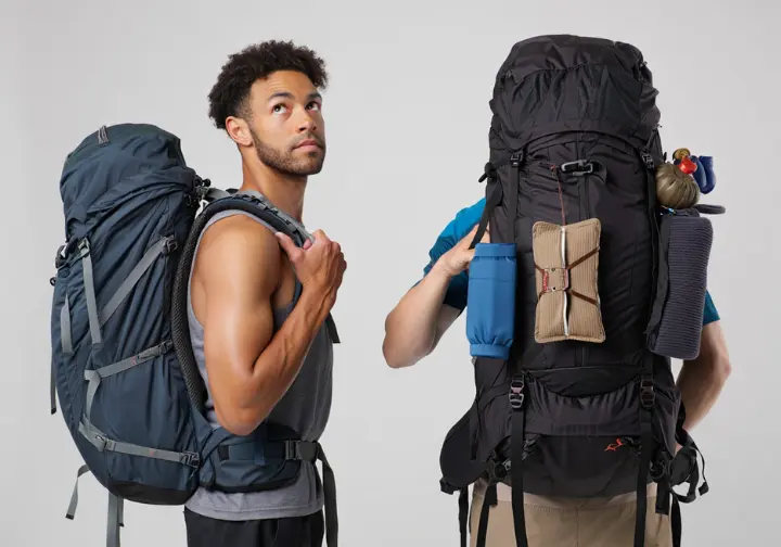 A hiker stands thoughtfully between a small ultralight backpack and a larger traditional backpack, illustrating the philosophy of packing choice.