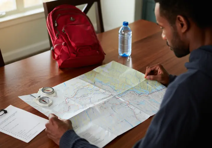 A hiker sits at a wooden table planning a trip to Turkey Mountain Nature Preserve with a map, compass, and day-hiking gear.