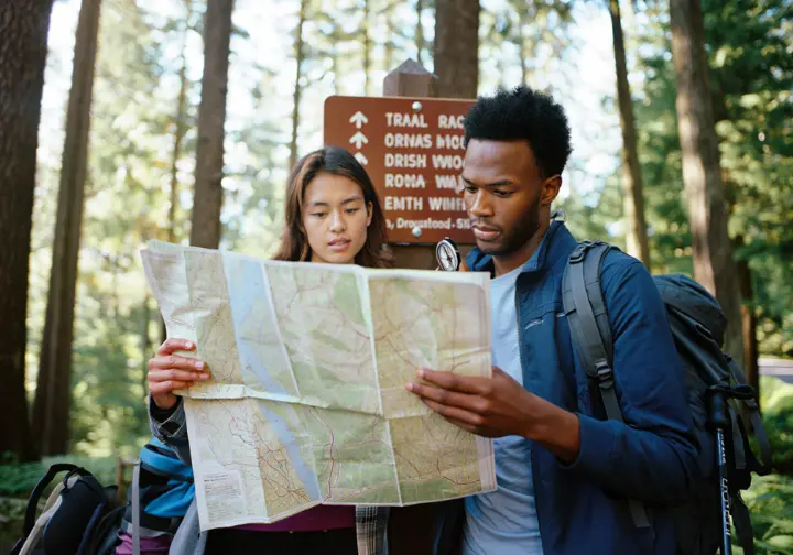Two hikers carefully planning their route with a map and compass at a trailhead, demonstrating hiking safety and preparation.