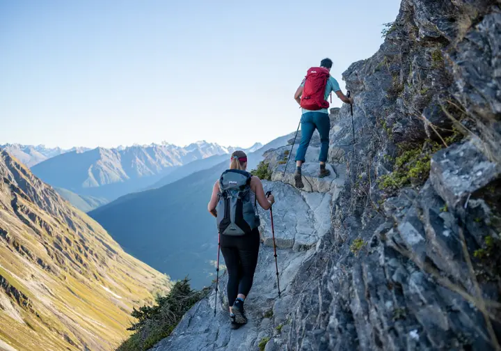 Two backpackers carefully hiking on a narrow, exposed high-alpine trail in Glacier National Park with panoramic mountain views.