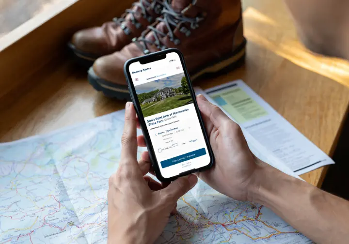 A hiker plans their trip, holding a smartphone showing a confirmed parking reservation for Sam's Point, with a trail map and boots on the table.