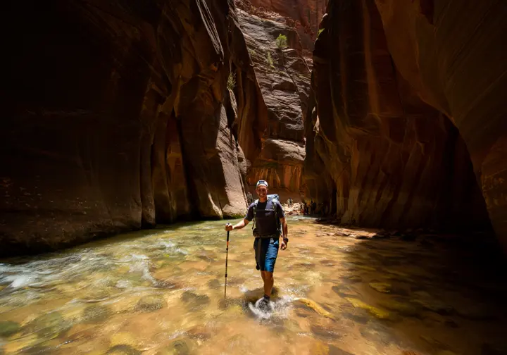A hiker uses trekking poles for balance while carefully wading through a river in a narrow slot canyon, demonstrating a skill required for technical day hikes.