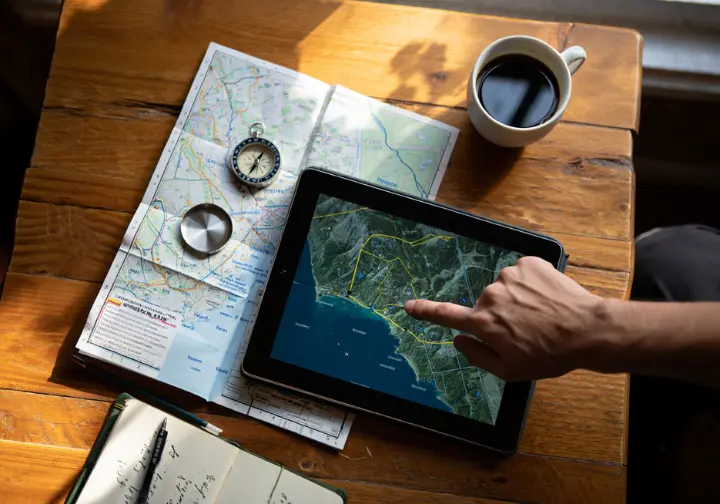 A hiker compares trail options for a Vancouver hike using a tablet, a physical map, and a compass on a wooden table.