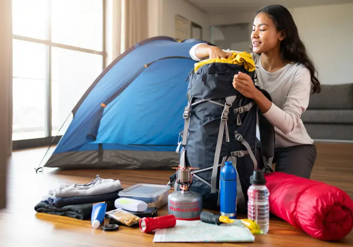 A female beginner hiker kneels on her floor, packing her backpack for her first overnight trip with essential gear laid out around her.