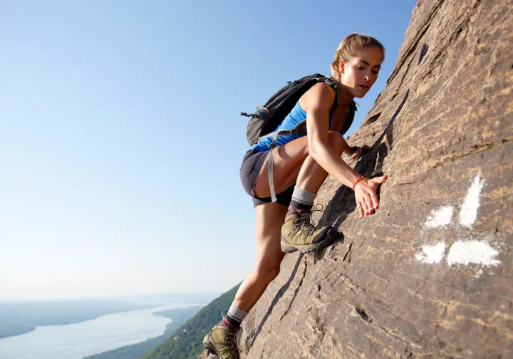 A female hiker confidently rock scrambling up the steep Breakneck Ridge trail, with the Hudson River visible below.