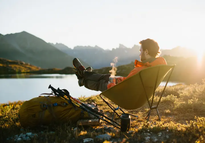 A hiker relaxes in a lightweight camp chair at a scenic campsite, demonstrating the comfort upgrades for an intermediate backpacker.