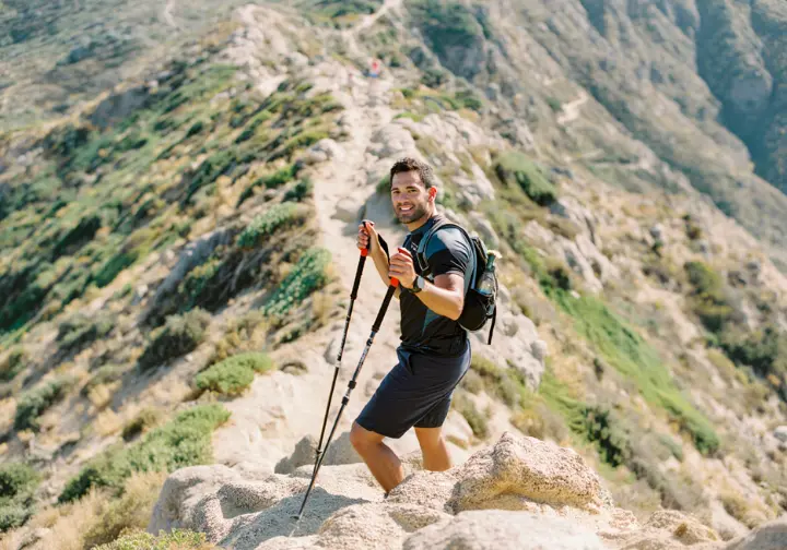 A hiker using trekking poles pauses on a switchback to look down a moderate mountain trail, showcasing the process of honing his hiking craft.