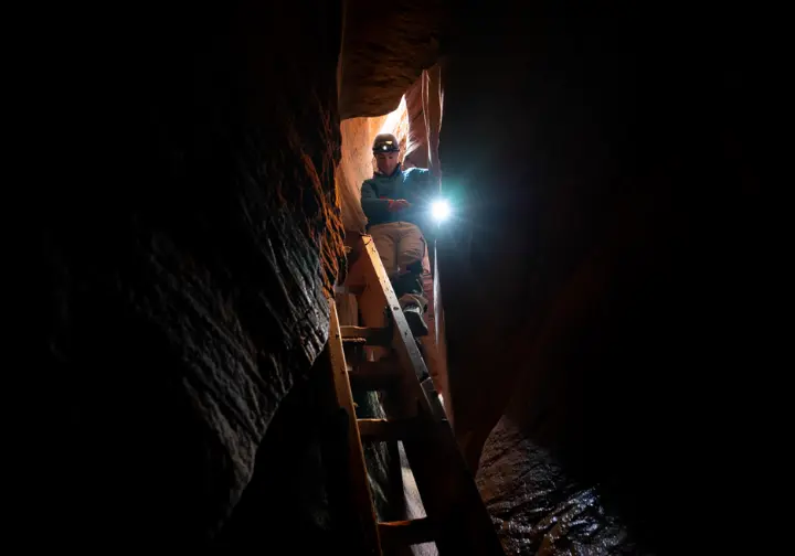 A hiker with a headlamp climbs a wooden ladder inside the dark, narrow rock crevices of the famous Ice Caves landmark at Sam's Point Preserve.