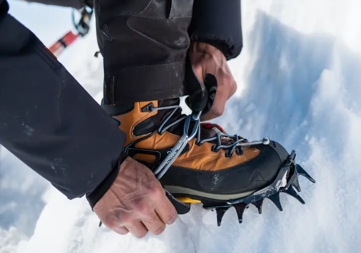 Close-up of a hiker's hands attaching crampons to mountaineering boots in a snowy, advanced alpine environment.