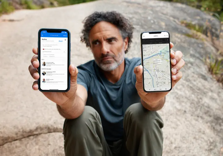 An experienced hiker sits on a rock comparing two smartphones, one showing a trail discovery app and the other a professional navigation app, demonstrating the two-app strategy.