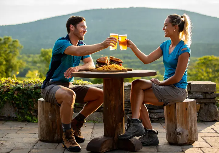 Two hikers celebrate after their Mohonk adventure at a restaurant in New Paltz, with the Shawangunk mountains in the background.