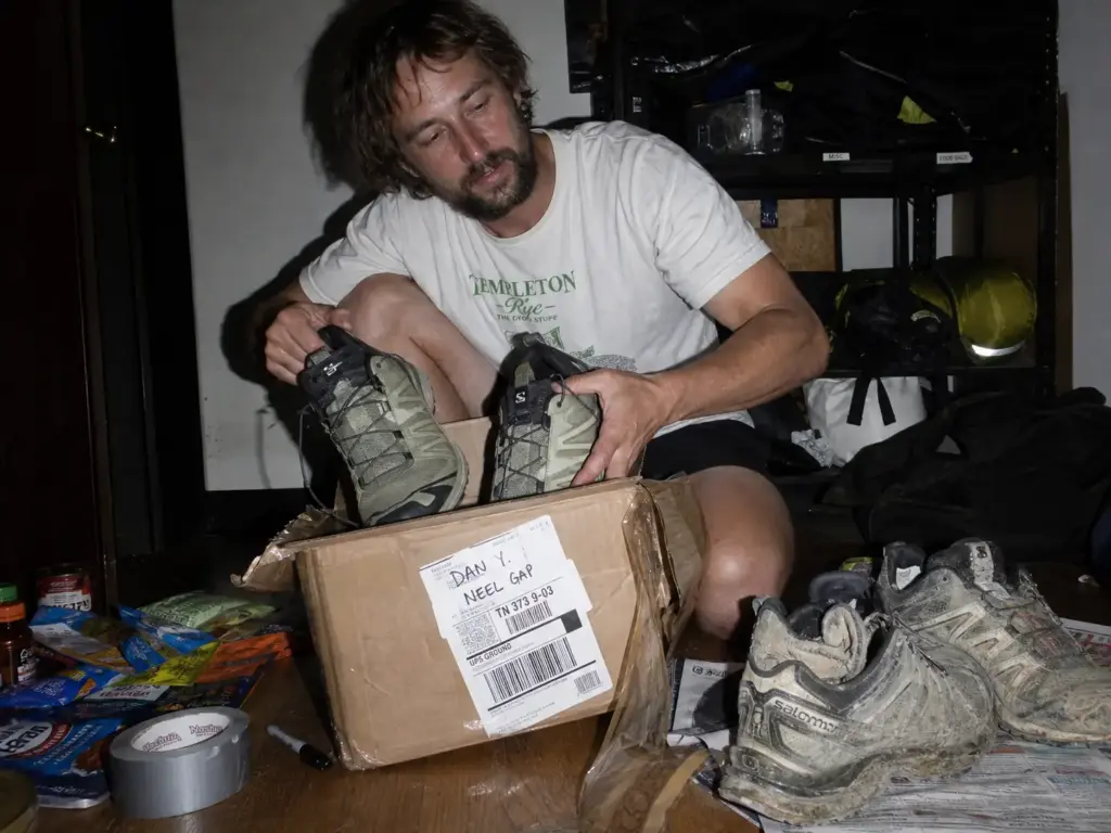 Thru-hiker opening a mailed resupply box with a fresh pair of trail runners in a trail town