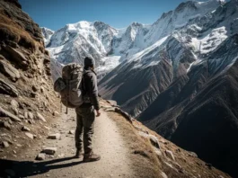 A full-body shot of a solo hiker with a backpack standing on a high-altitude trail overlooking a vast mountain range.