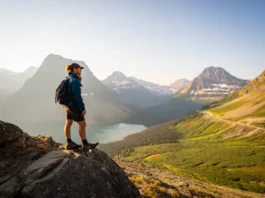 A hiker stands on a scenic overlook on the Highline Trail, thoughtfully planning their route while looking out over the mountains and valleys of Glacier National Park at sunset.