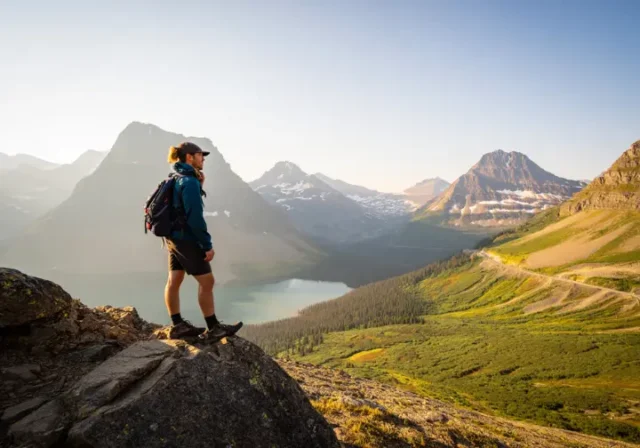 00 glacier-np-day-hikes-planning-guide A hiker stands on a scenic overlook on the Highline Trail, thoughtfully planning their route while looking out over the mountains and valleys of Glacier National Park at sunset.