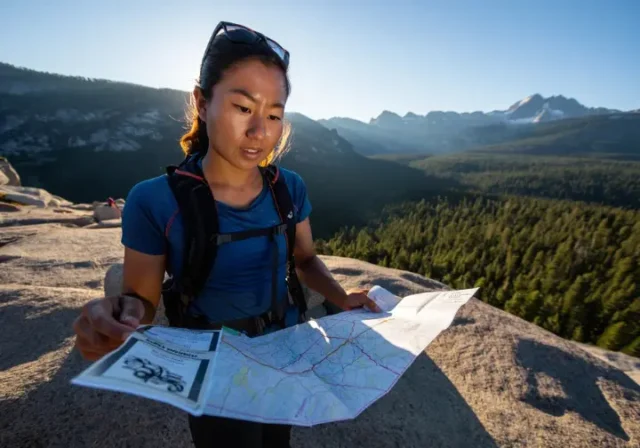 A female hiker stands on a granite ridge overlooking a vast Northern California landscape, carefully planning her route using a map and permit.