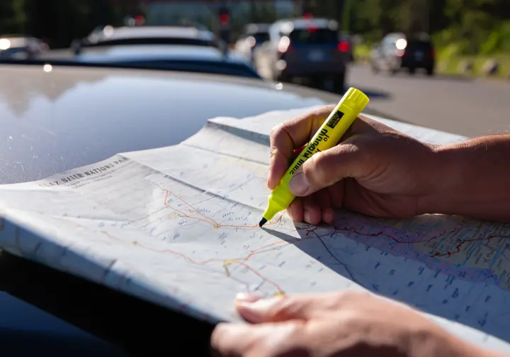 A hiker's hands trace a route on a Glacier National Park map spread on a car hood, with a line of cars at a park entrance visible in the background, illustrating the importance of logistical planning.