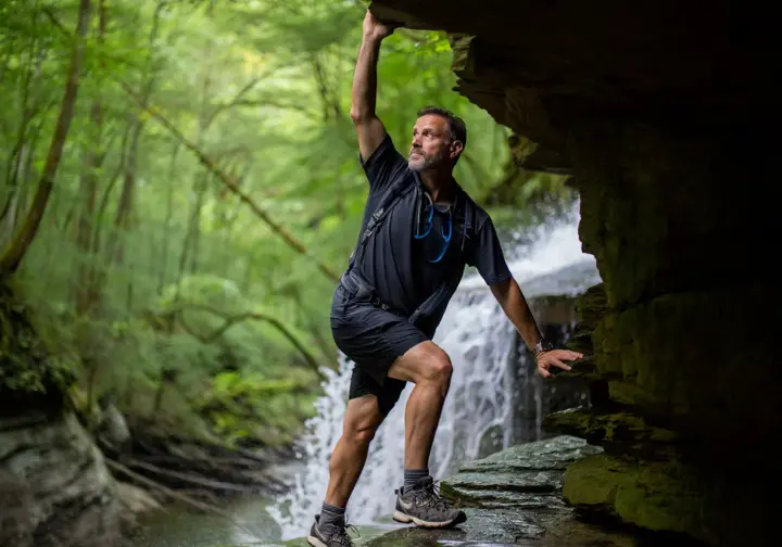 A hiker carefully navigates the wet, mossy stone steps of the Falls Trail at Ricketts Glen State Park, with a cascading waterfall in the background.