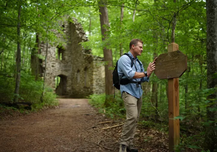 A hiker pauses on a forested trail to read a historical marker, with the stone ruins of the Center Furnace visible in the background at Land Between the Lakes.