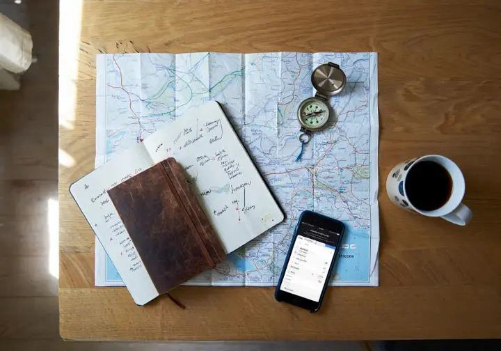 A flat lay of hiking trip planning gear for the Lofoten Islands, including a map, compass, notebook, and smartphone showing a weather forecast.