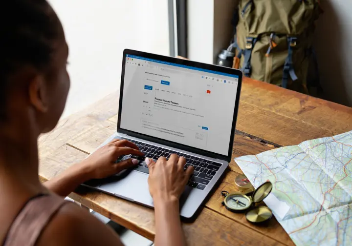 A hiker carefully plans her Colorado backpacking trip on a laptop, viewing the permit reservation system with a map and compass nearby.