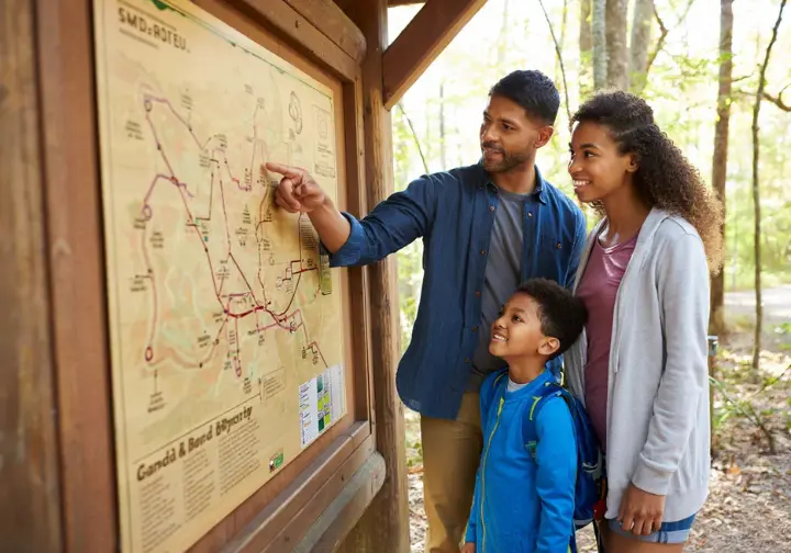 A diverse family of hikers stands together at a trailhead kiosk, pointing at a large park map to choose the perfect trail for their day's goal.