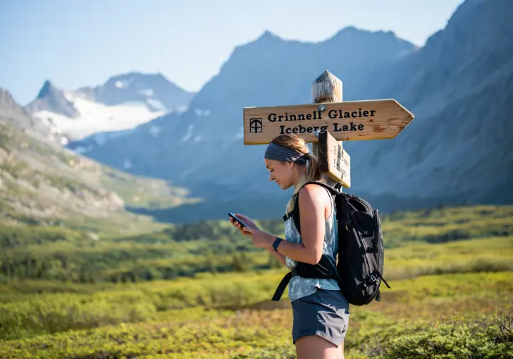 A hiker standing at a trail junction sign in Glacier National Park, deciding between the Grinnell Glacier and Iceberg Lake trails, representing the choice of the best hike experience.