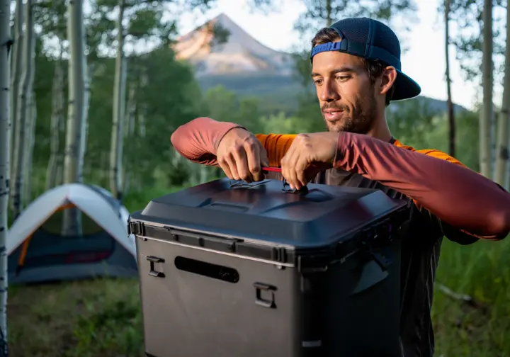 A backpacker demonstrates a critical Colorado-specific safety protocol by properly locking a hard-sided bear canister at his campsite.