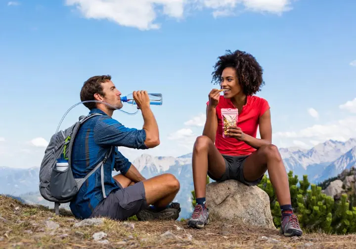 Two hikers fueling their hike with water from a hydration reservoir and eating trail mix during a break on a scenic trail.