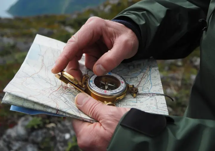 A close-up of a hiker's hands using a compass and a topographic map to navigate safely on a trail in the Lofoten Islands.