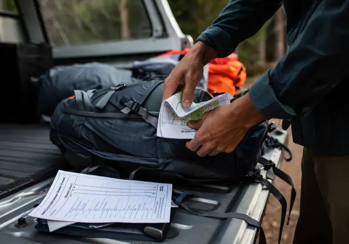 A hiker efficiently packs a backpack at their car's tailgate, with a non-quota wilderness permit and a map of the Trinity Alps laid out, ready for a last-minute trip.