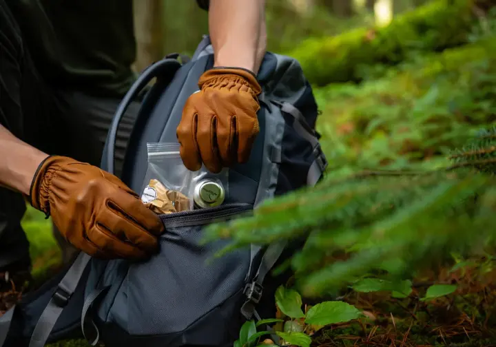 A close-up of a hiker's hands placing a small bag of collected litter into their backpack, demonstrating responsible Leave No Trace practices on the trail.