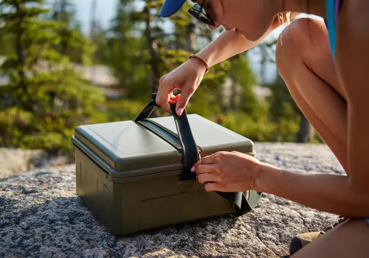 A close-up of a hiker's hands correctly locking a mandatory hard-sided bear canister at a campsite, demonstrating an essential backcountry best practice.
